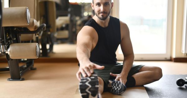 A man in gym clothes stretches on the floor in a modern fitness center.