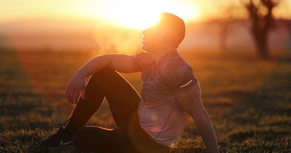Muscular man sits in rural field at sunset, exhaling visible breath.