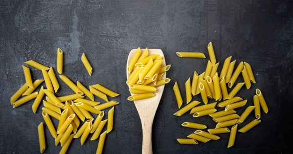 Top view of uncooked penne pasta and wooden spoon on a black surface, perfect for food-related content.