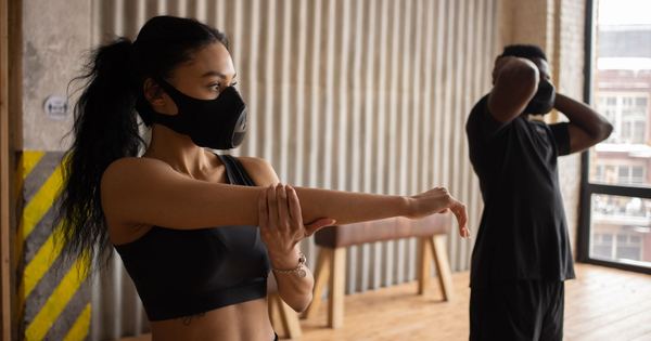 African American sportswoman in textile mask stretching arm against anonymous male partner with hands behind head working out in gymnasium