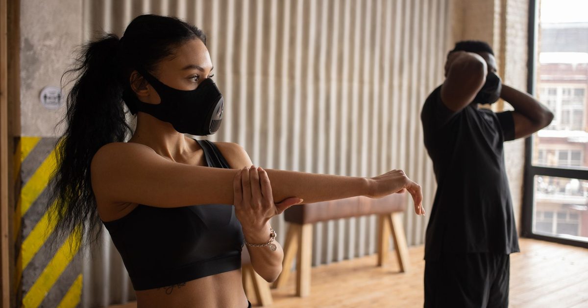 African American sportswoman in textile mask stretching arm against anonymous male partner with hands behind head working out in gymnasium