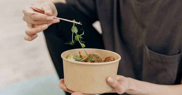 A woman holding a paper bowl filled with a healthy vegetarian meal in an indoor setting, promoting wellness.